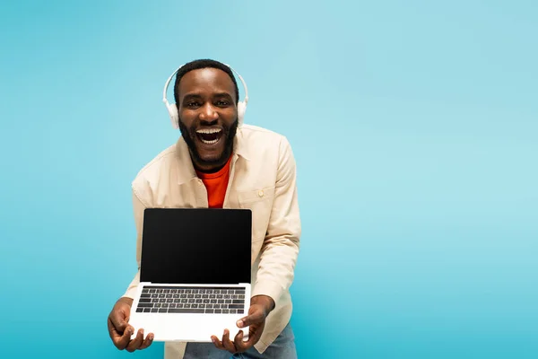 Homme afro-américain étonné dans les écouteurs montrant ordinateur portable avec écran vide isolé sur bleu — Photo de stock