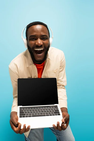 Rindo afro-americano homem em fones de ouvido mostrando laptop com tela em branco no azul — Fotografia de Stock