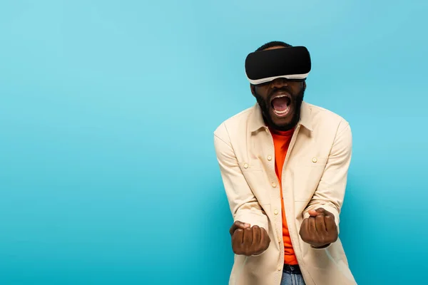 Homme afro-américain en colère dans le casque vr criant et montrant les poings serrés isolés sur bleu — Photo de stock