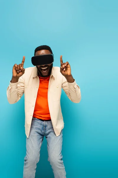 Amazed african american man in vr headset pointing up with fingers on blue background — Stock Photo