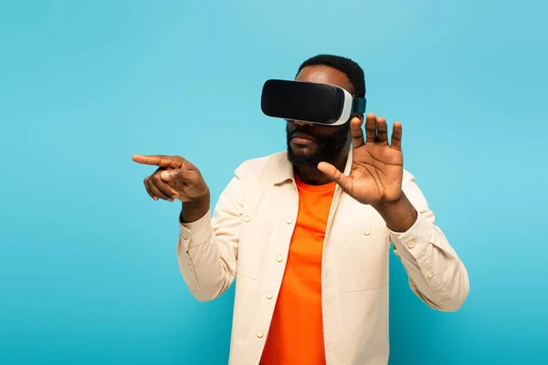 Hombre afroamericano en auriculares vr apuntando con el dedo mientras toca algo aislado en azul - foto de stock