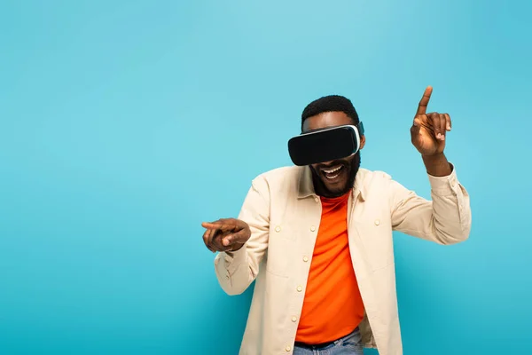 Cheerful african american man in vr headset pointing with fingers isolated on blue — Stock Photo