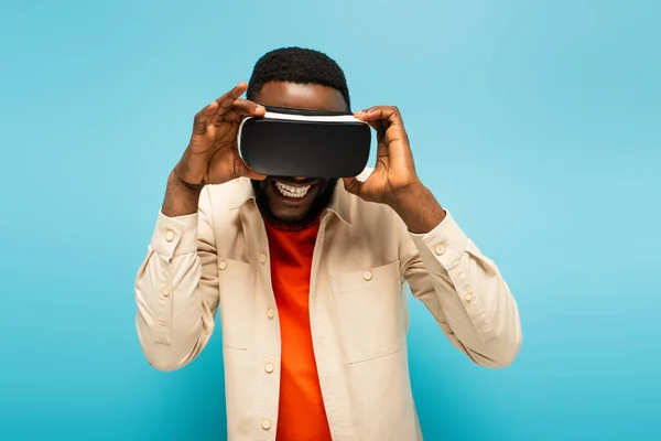 Улыбающийся африканский американец трогает наушники VR, в то время как игры изолированы на синий — стоковое фото