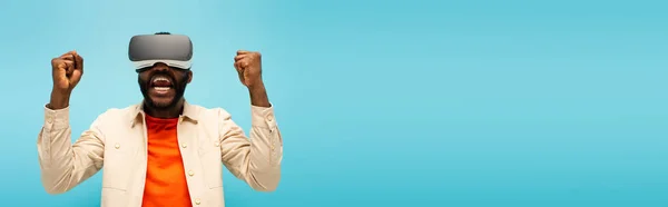 Hombre afroamericano emocionado mostrando gesto de victoria mientras juega en auriculares vr aislados en azul, bandera - foto de stock