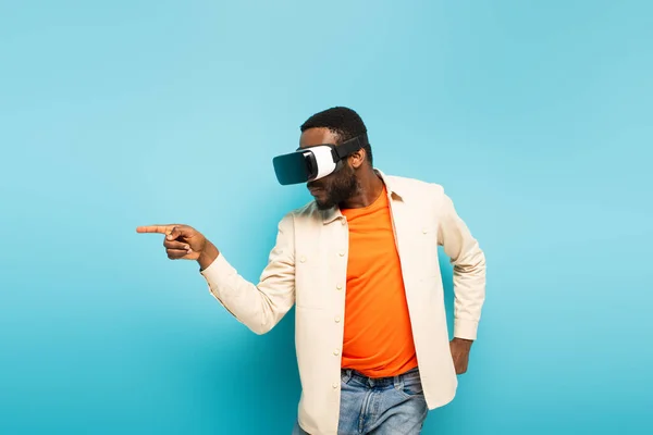 Young african american man pointing aside with finger while gaming in vr headset on blue background — Stock Photo