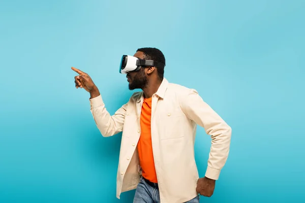 Африканский мужчина показывает пальцем во время игры в гарнитуру VR на синем фоне — стоковое фото