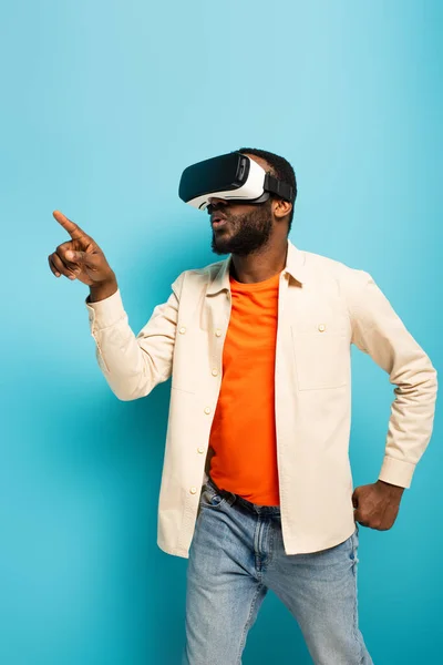 African american man in vr headset pointing away with finger on blue background — Stock Photo