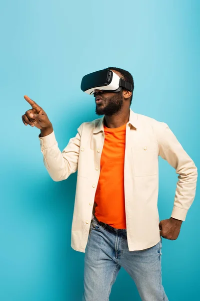 Africano americano hombre en vr auriculares apuntando lejos con dedo sobre fondo azul - foto de stock