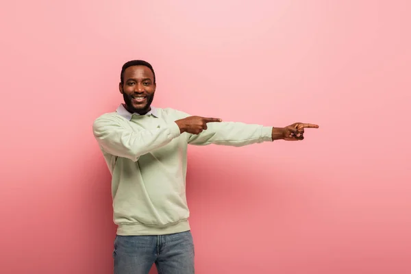 Hombre afroamericano feliz apuntando lejos con los dedos en el fondo rosa - foto de stock