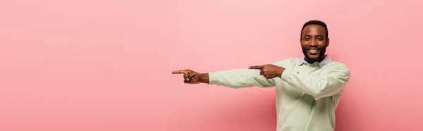 Homme afro-américain joyeux pointant de côté avec les doigts sur fond rose, bannière — Photo de stock