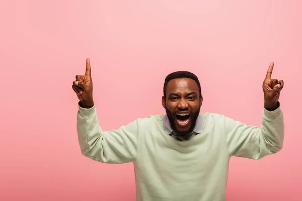 Surpreso afro-americano homem olhando para a câmera enquanto apontando para cima com os dedos isolados em rosa — Stock Photo