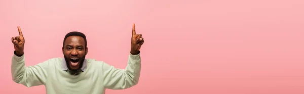 Thrilled african american man with open mouth pointing up with fingers isolated on pink, banner — Stock Photo