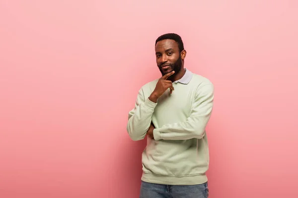 Thoughtful african american man looking at camera while holding hand near face on pink background — Stock Photo