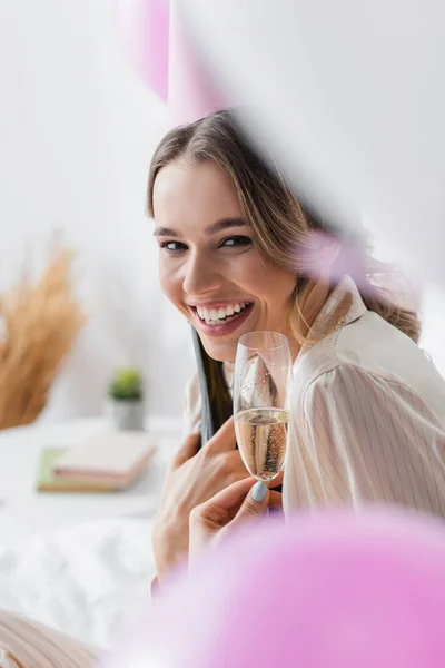 Donna sorridente nel cappuccio del partito che tiene il vetro di champagne vicino ai palloncini sfocati — Foto stock