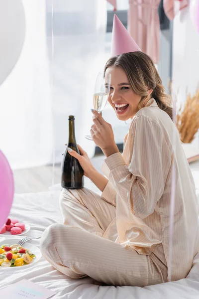 Cheerful woman with champagne celebrating birthday near fruit salad and balloons in bedroom — Stock Photo