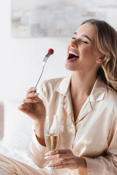 Positive Frau im Pyjama hält Champagner und Himbeere auf Gabel im Schlafzimmer — Stockfoto