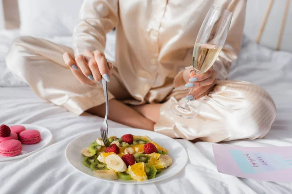 Cropped view of woman with champagne holding fork near fruit salad, macarons and birthday greeting card on bed — Stock Photo