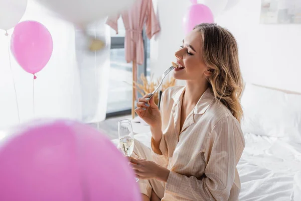 Mujer sonriente con champagne oyendo plátano cerca de globos en el dormitorio - foto de stock