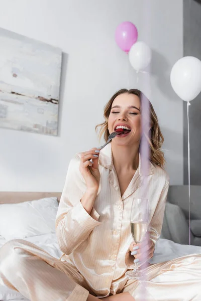 Positive Frau mit Glas Champagner isst Himbeere in der Nähe von Luftballons im Schlafzimmer — Stockfoto