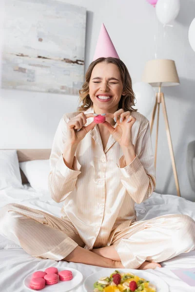 Positive woman in party cap holding macaroon near fruit salad and balloons in bedroom — Stock Photo