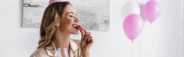 Smiling woman in party cap holding glass and eating macaroon at home, banner — Stock Photo