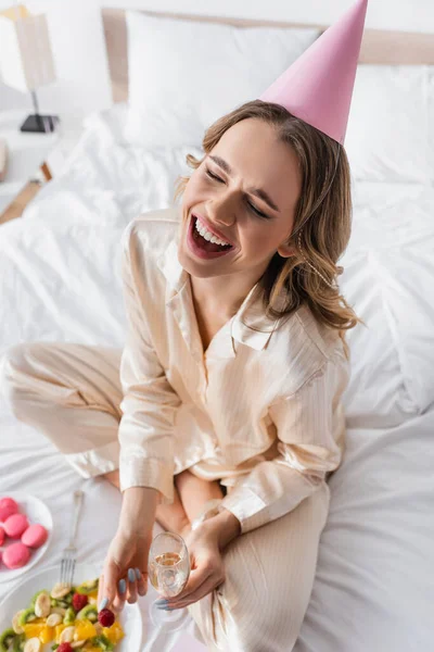 Cheerful woman in party cap holding champagne and raspberry on bed — Stock Photo