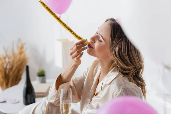 Mujer joven con champán soplando cuerno de fiesta cerca de globos - foto de stock