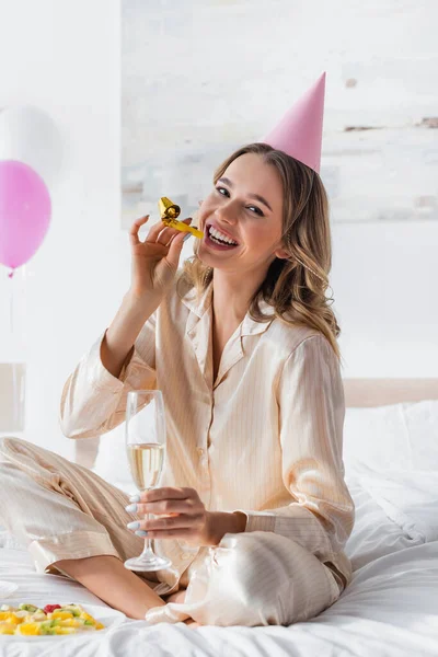 Femme souriante avec champagne tenant corne de fête près de salade de fruits sur le lit — Photo de stock