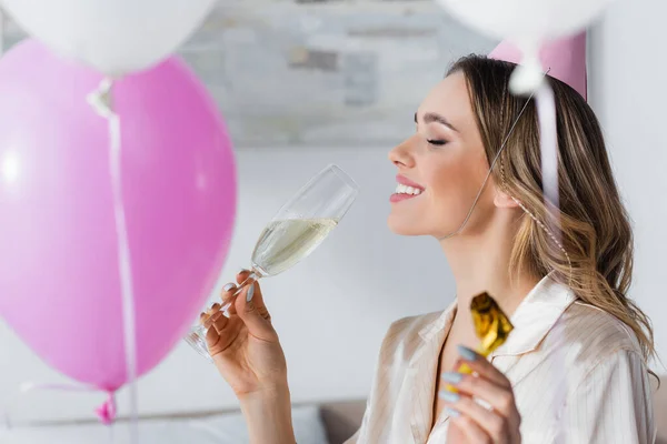 Vista lateral de una mujer sonriente sosteniendo champán y cuerno borroso cerca de globos - foto de stock