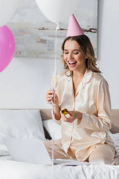 Mulher sorridente no pijama segurando champanhe e chifre de festa perto do laptop na cama — Fotografia de Stock