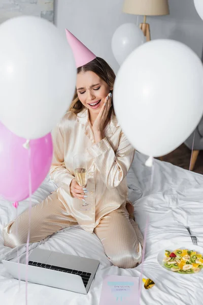 Woman with champagne having video call on laptop near greeting card, balloons and fruit salad in bedroom — Stock Photo