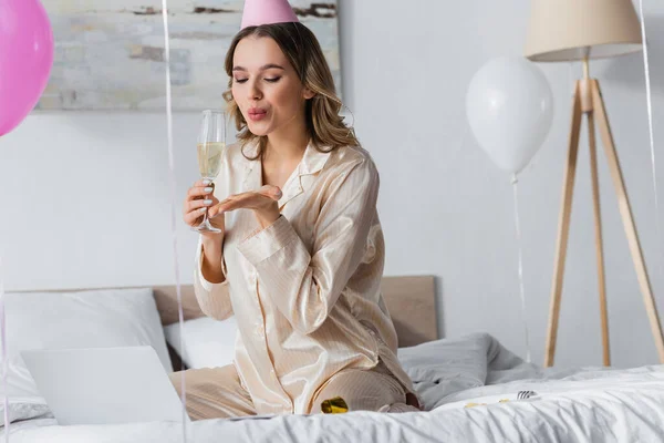 Frau mit Champagner pustet Luftkuss während Videoanruf auf Laptop auf Bett — Stockfoto