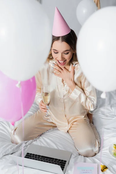 Fröhliche Frau mit Champagner beim Videochat auf Laptop in der Nähe von Geburtstagsgrußkarte auf dem Bett — Stockfoto