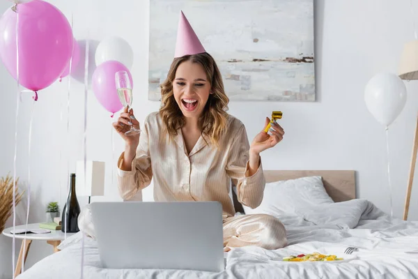 Donna positiva con corno del partito e champagne che fa videochiamata sul computer portatile mentre festeggia il compleanno in camera da letto — Foto stock