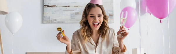 Donna felice con corno partito e champagne che festeggia il compleanno vicino ai palloncini a casa, striscione — Foto stock