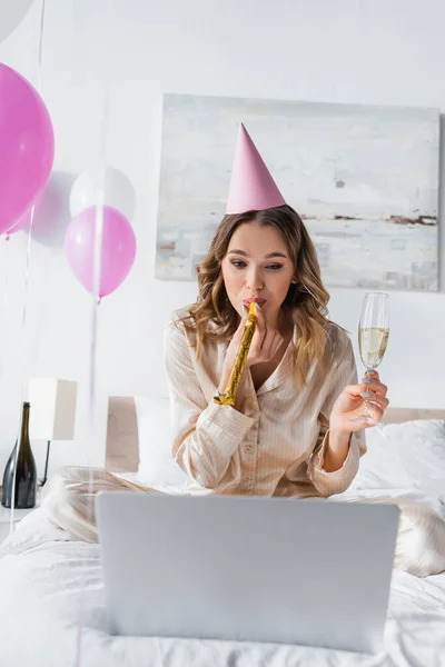 Woman with champagne blowing party horn near blurred laptop on bed — Stock Photo