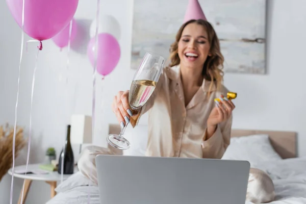 Шампанское в руке размытой женщины, празднующей день рождения возле ноутбука на кровати — стоковое фото