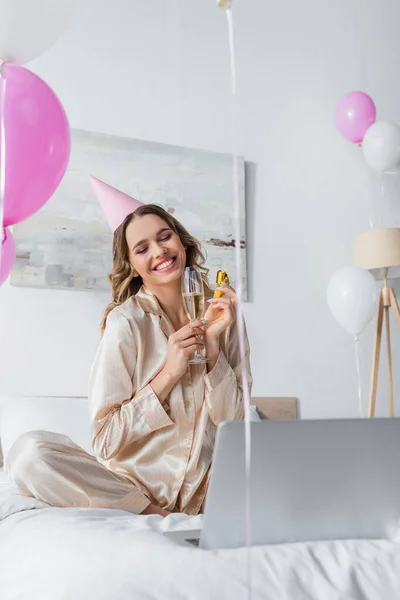 Happy woman with party blower, champagne and cap having video call on laptop during birthday — Stock Photo