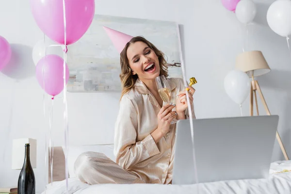 Mulher comemorando aniversário com champanhe e chifre de festa enquanto tem videochamada no laptop — Fotografia de Stock