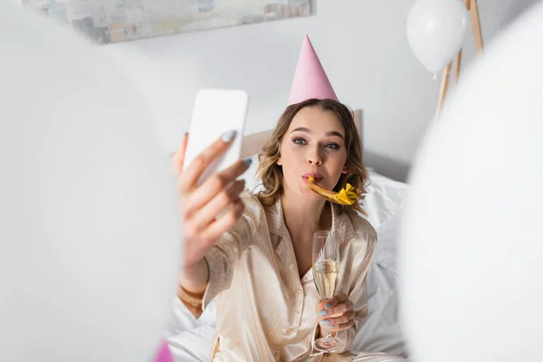 Mulher com chifre de festa e champanhe tirando selfie durante festa de aniversário no quarto — Fotografia de Stock