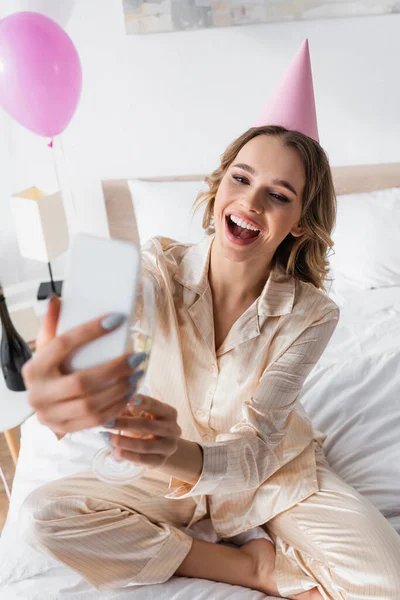 Donna positiva con champagne e cappuccio partito avendo videochiamata su smartphone in camera da letto — Foto stock