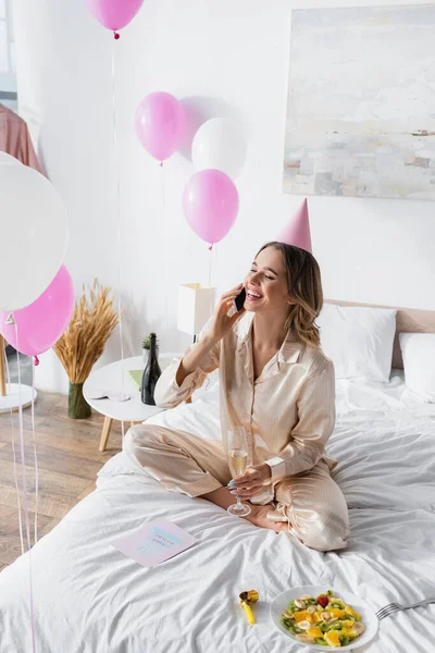 Mulher feliz falando no smartphone enquanto comemora o aniversário com champanhe no quarto — Fotografia de Stock