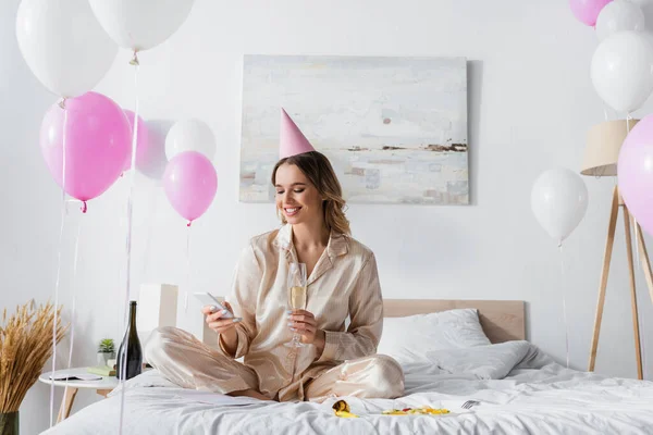 Femme souriante avec champagne à l'aide d'un smartphone près de la salade de fruits et ballons dans la chambre — Photo de stock