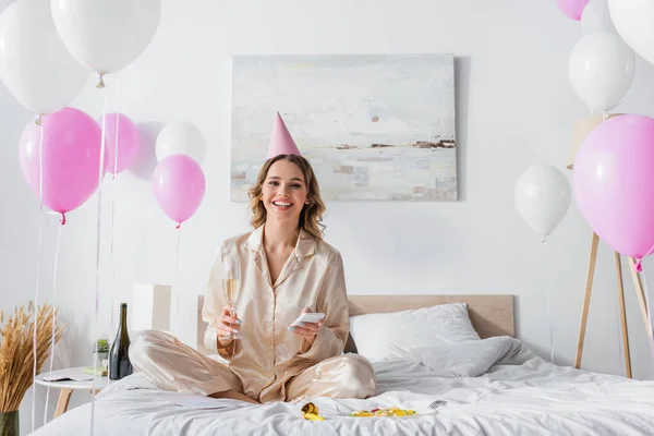 Mujer sonriente con teléfono inteligente y champán celebrando cumpleaños en el dormitorio - foto de stock