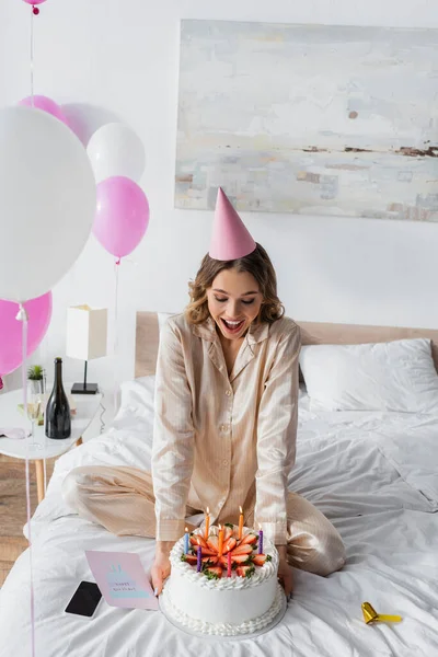 Donna eccitata in corno partito in possesso di torta di compleanno vicino biglietto di auguri e smartphone — Foto stock