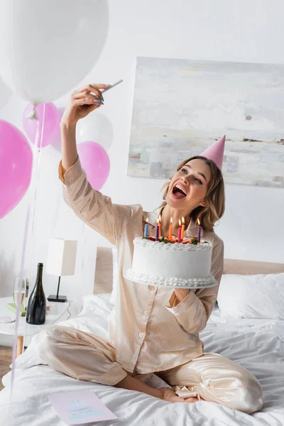 Excited woman in party cap taking selfie with smartphone and holding birthday cake — Stock Photo