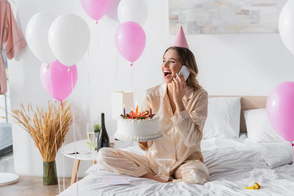 Aufgeregte Frau telefoniert und hält Geburtstagstorte mit Kerzen auf dem Bett — Stockfoto