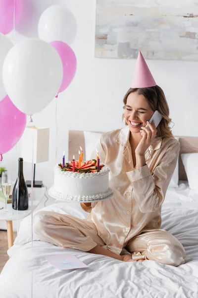 Fröhliche Frau telefoniert mit Handy und hält Geburtstagstorte bei Feier im Schlafzimmer — Stockfoto