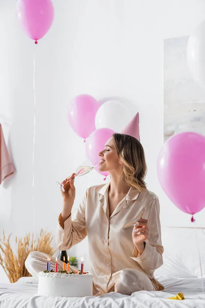 Vista lateral de la mujer sonriente bebiendo champán cerca de la torta con velas durante la celebración del cumpleaños - foto de stock