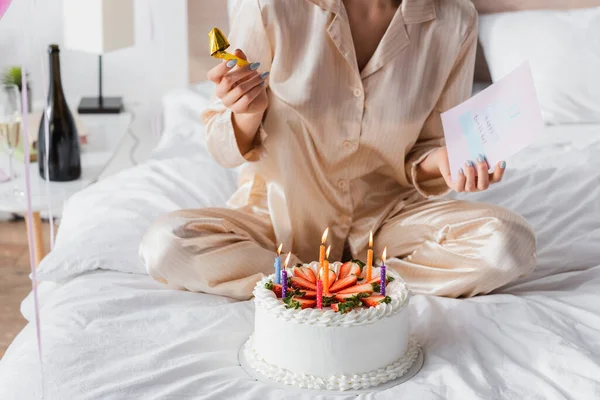 Vista cortada da mulher no pijama segurando chifre do partido e cartão de saudação perto do bolo de aniversário na cama — Fotografia de Stock